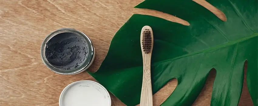 A close-up of a jar of charcoal toothpaste, a toothbrush, and a green leaf on a wooden surface.