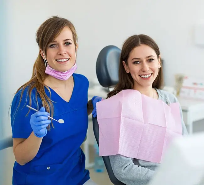 A close-up of a smiling dentist in a blue lab gown holding a mouth mirror in her hands stands beside a smiling patient sitting on an Orthodontic chair.
