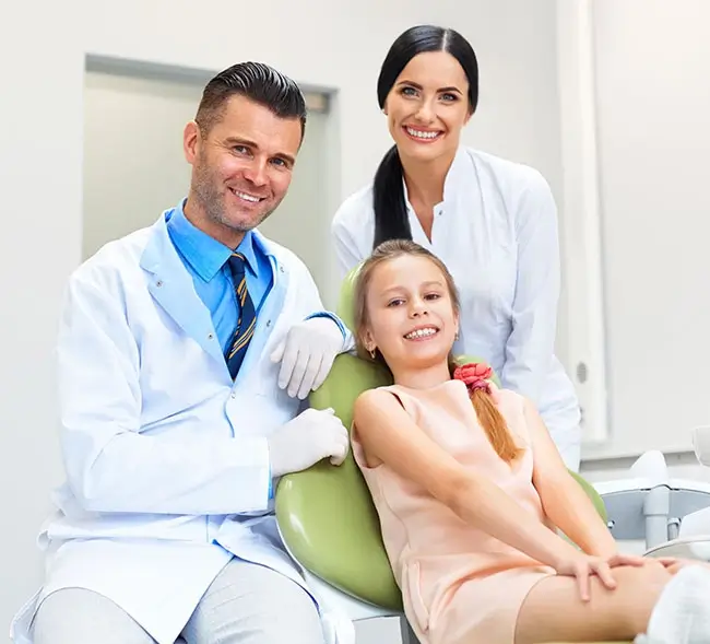 A full shot of a team of two dentists with a smiling little girl in a pink dress inside a clinic.