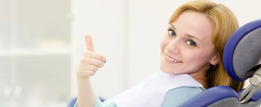 A close-up of a smiling blond woman sitting on an Orthodontic chair doing a thumbs-up gesture.