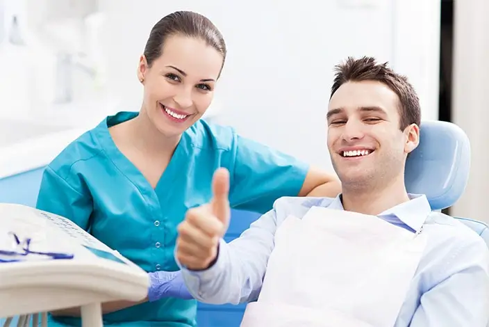 A close-up of a man doing a thumbs-up gesture sitting on an Orthodontic chair with a nurse in a teal lab gown sitting beside him.