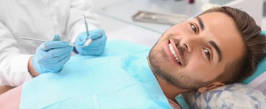A close-up of a smiling man laying on an Orthodontic chair and a hand holding a dental instrument.