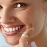 A close-up of a smiling woman holding dental floss near his mouth.
