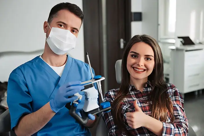 A close-up of a dentist in a blue lab gown wearing a mask and a smiling patient in a checkered polo doing a thumbs-up gesture.