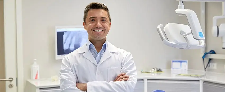 A close-up of a smiling dentist wearing a white robe inside a dental clinic.